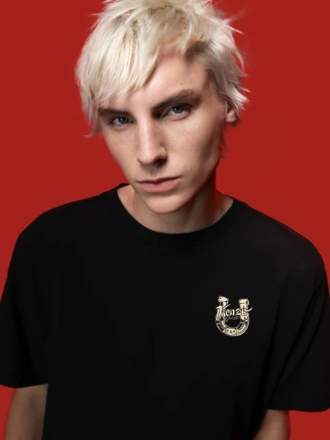 Portrait of a young adult male with platinum blond hair posing in front of a red backdrop while wearing a black t-shirt.