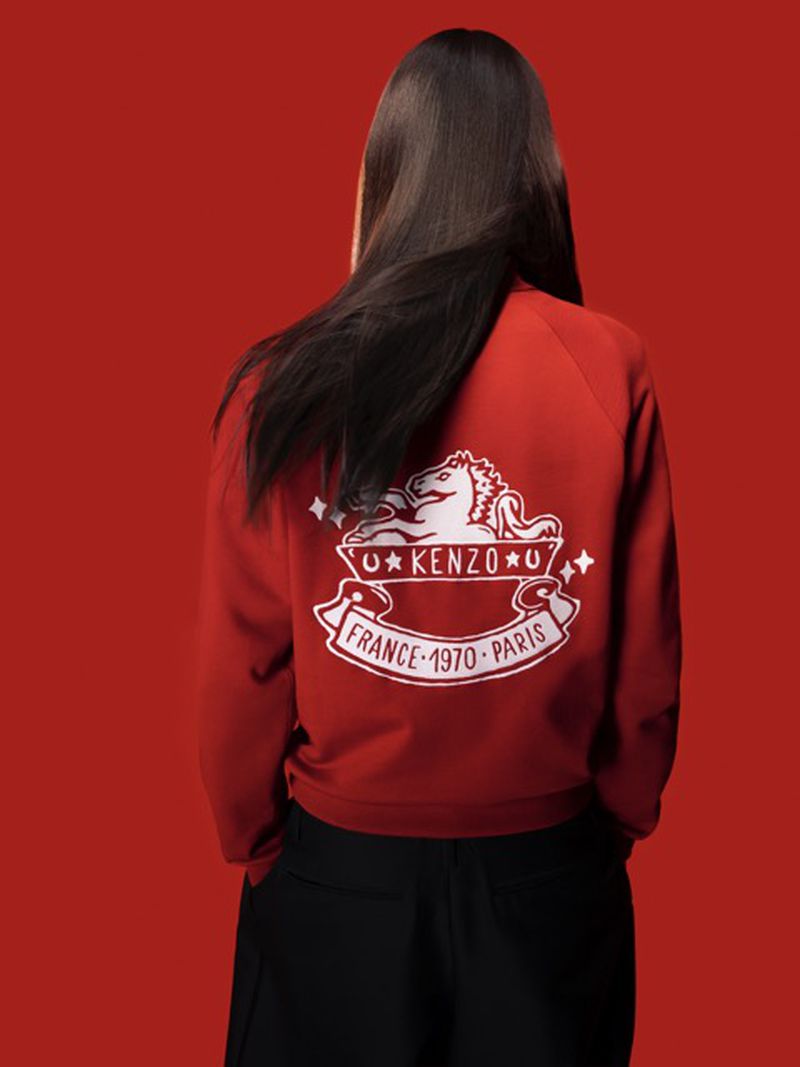 Back view of a woman with long brunette hair wearing a Kenzo logo sweatshirt against a red background in a studio photograph.