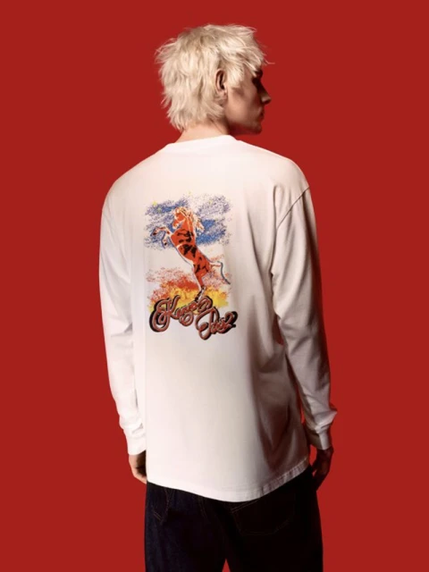 An adult male with light blonde hair is shown from the back, wearing a white long-sleeve shirt with a graphic design, against a red background.