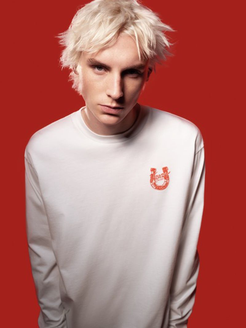 Close-up portrait of a young adult with pale skin, light blonde hair, wearing a white long-sleeved shirt with a small graphic on a red background.