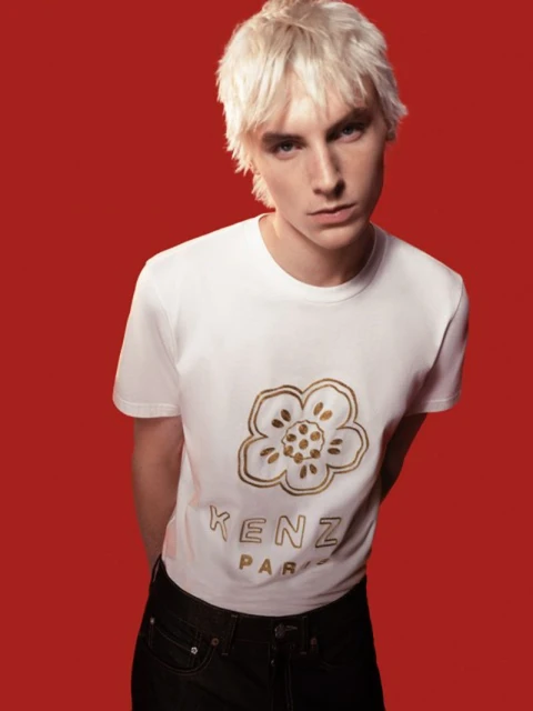 Blonde male model in a white Kenzo t-shirt poses against a red backdrop, shot at eye-level in a fashion editorial style, studio lighting.