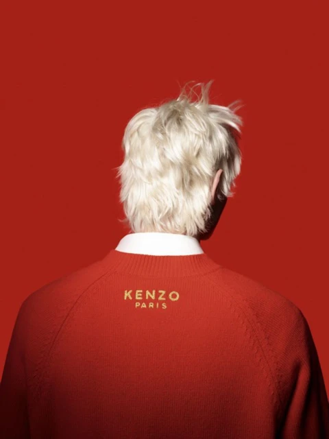From behind, a fair-skinned person with blond hair wears a red Kenzo sweater against a matching red backdrop in a studio setting.