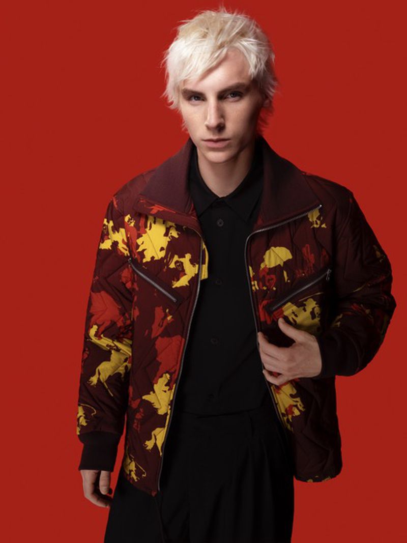 Studio shot of a male model with blonde hair wearing a patterned jacket, black shirt and pants against a solid red background.