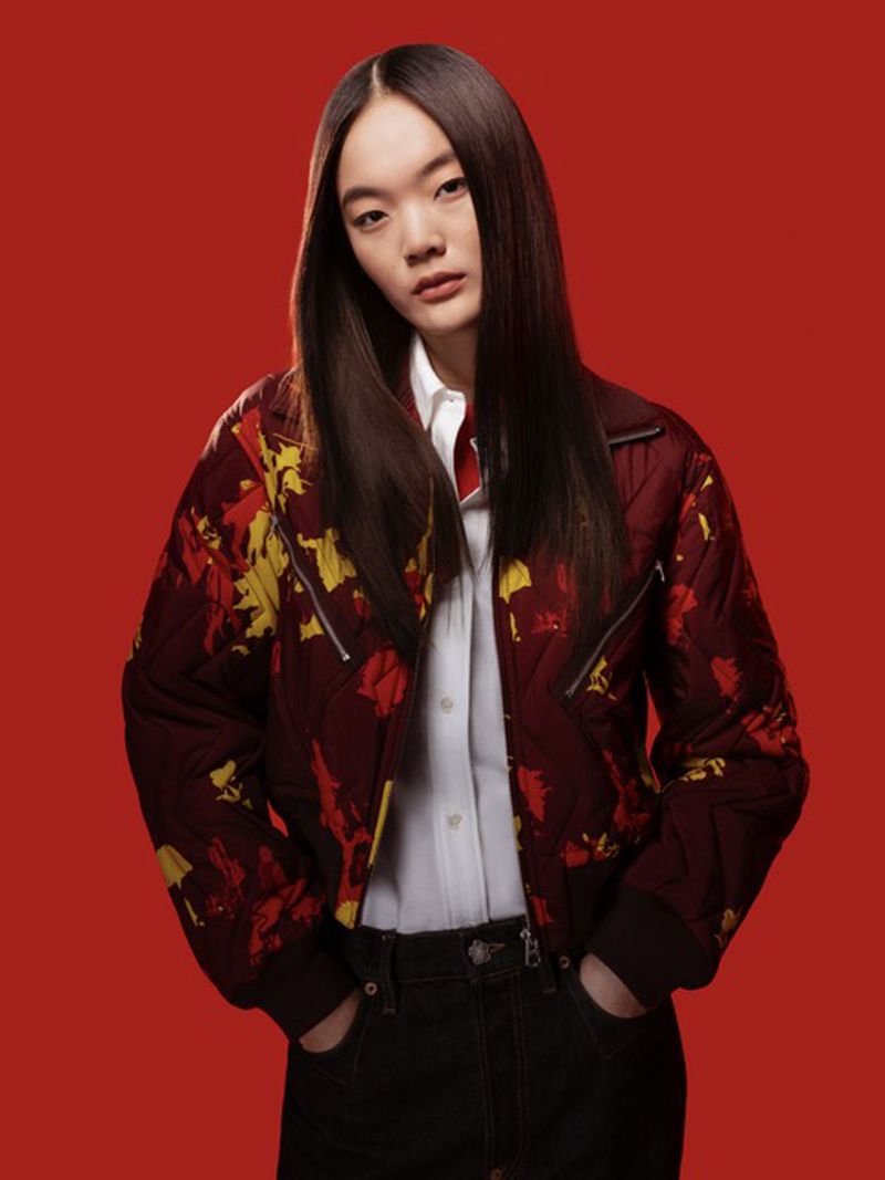 Portrait of an Asian female wearing a patterned zip-up jacket, white button-down shirt, and dark denim jeans against a red backdrop.