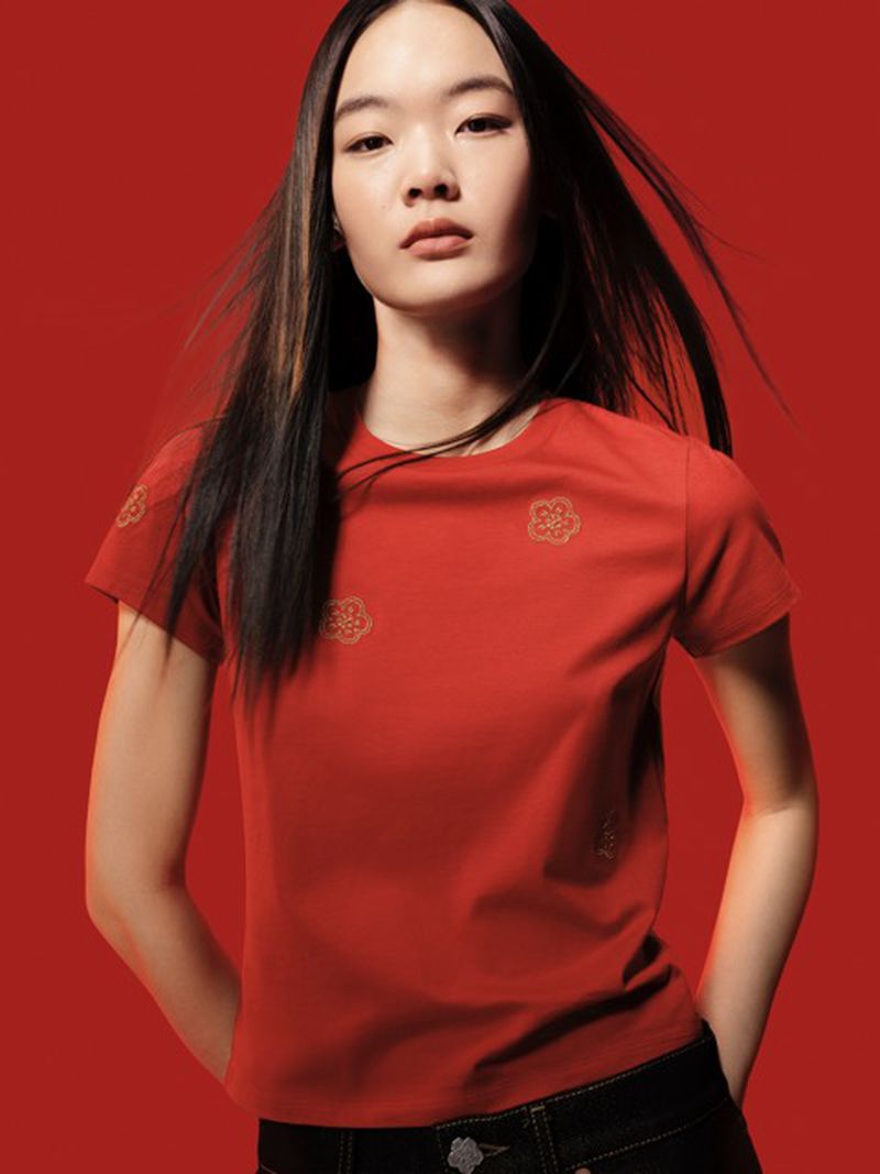 Fashion shot of a young Asian woman in a red shirt with flower details, wearing jeans against a red background in a studio setting.