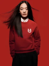 Model with windswept dark hair wearing a red Kenzo sweatshirt over a white collared shirt against a bold red studio backdrop.