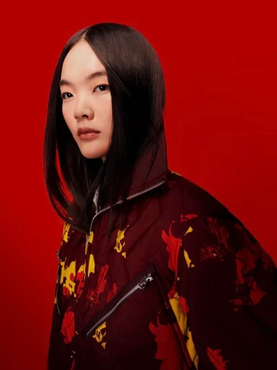Model Jiahui Meng wearing a printed maroon high-collar zip jacket against a solid red studio background for Kenzo.