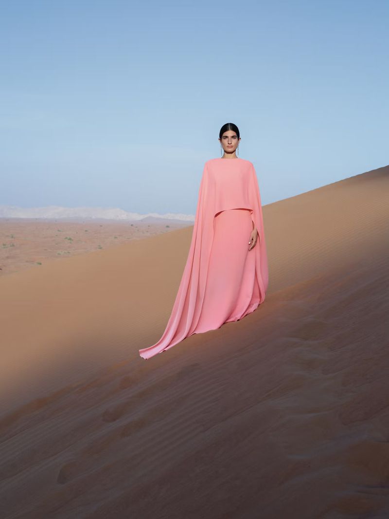 A woman in a flowing pink gown stands on a sand dune in a desert landscape under a clear sky, creating a striking fashion portrait.