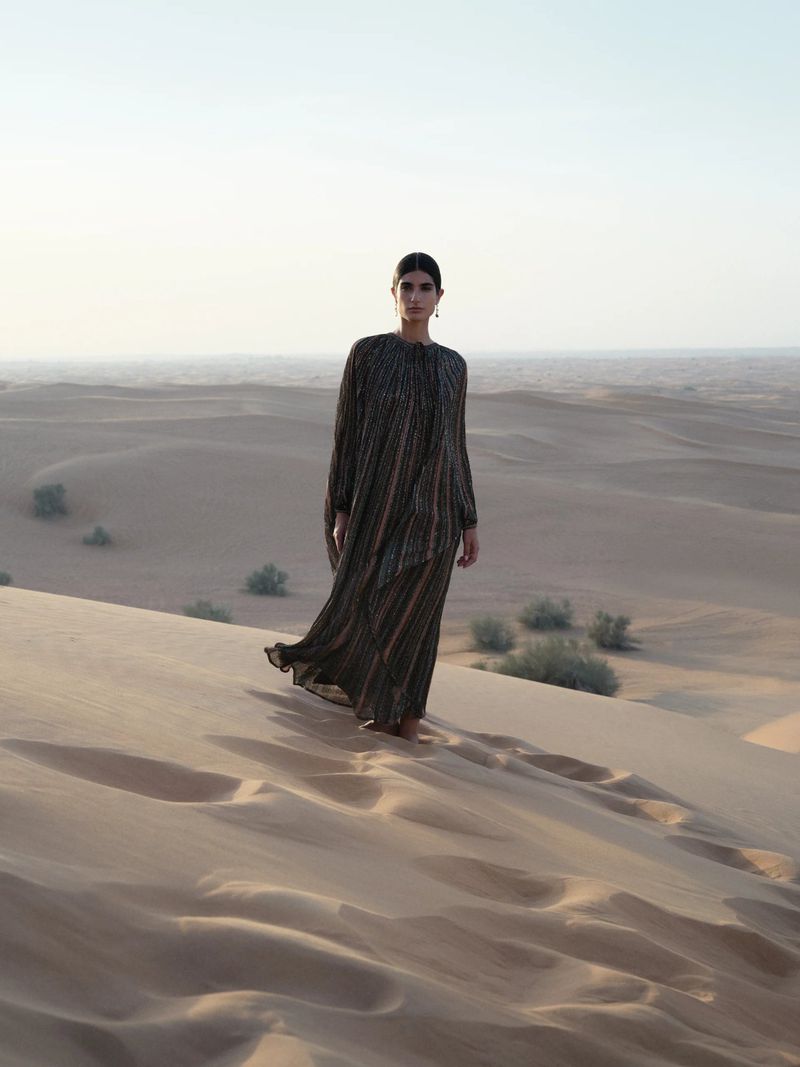 A woman stands on a sand dune in a flowing dress, creating a striking contrast against the desert landscape under a clear sky.