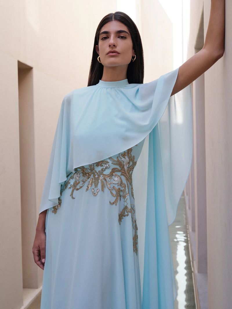 Elegant adult female in a light blue gown with gold embroidery, posing indoors with soft lighting and a neutral color palette.