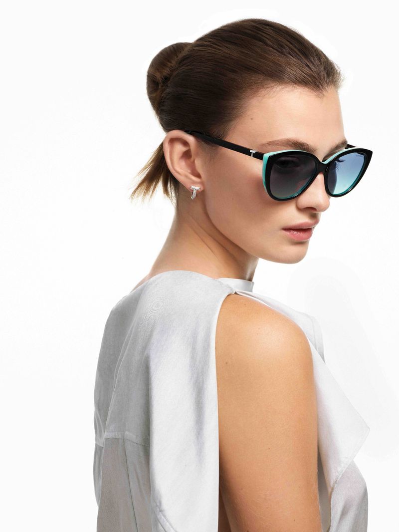 Portrait of a young woman wearing sunglasses and a light-grey top, posing against a white background in a fashion photography style.