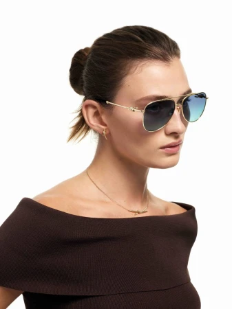 Portrait of a woman with brown hair wearing aviator sunglasses, a dark brown off-the-shoulder top, and a gold necklace against a white background.