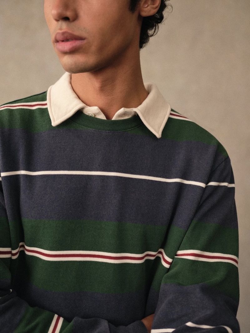 Close-up of a young adult man wearing a long-sleeved, horizontally striped rugby shirt in blue, green, and white against a neutral background.