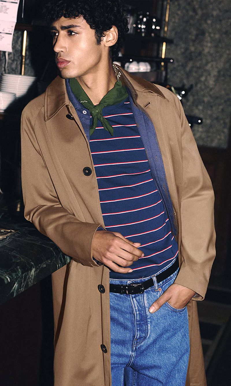 Handsome dark-haired man leans on a marble bar wearing a tan trench coat, striped shirt, jeans, and a green scarf.