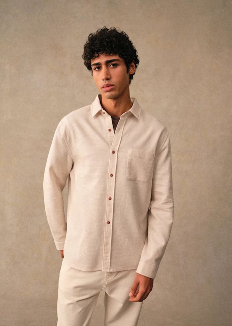 Model Maxime El Hanafi wearing a cream button-down overshirt and matching trousers against a neutral textured backdrop.