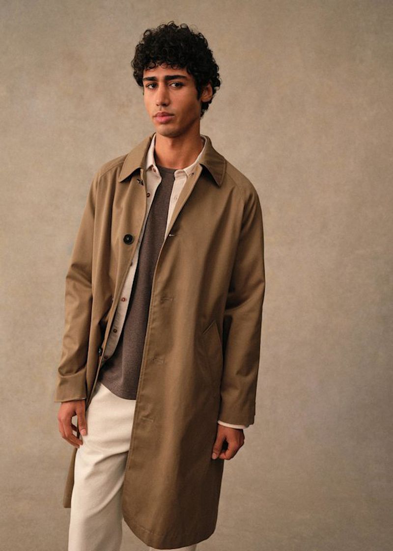 A curly-haired male model poses indoors in a brown trench coat, gray shirt, and white pants, set against a neutral backdrop in a studio setting.