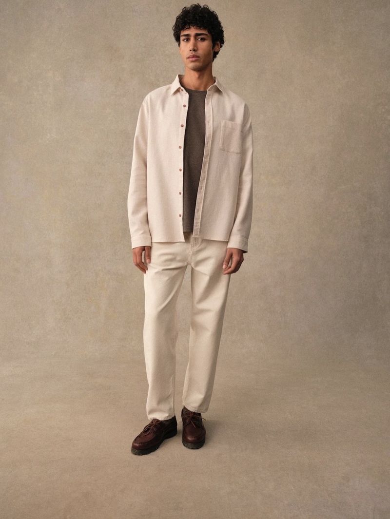 Full-shot of a young adult male with curly hair wearing an open beige shirt, t-shirt, beige pants, and dark brown shoes in studio setting.