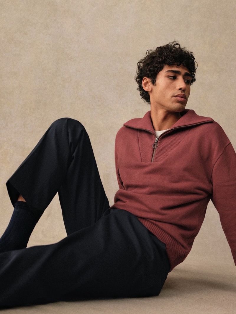 Male fashion model poses while seated indoors wearing a reddish-brown sweater and black trousers. Neutral-toned background.