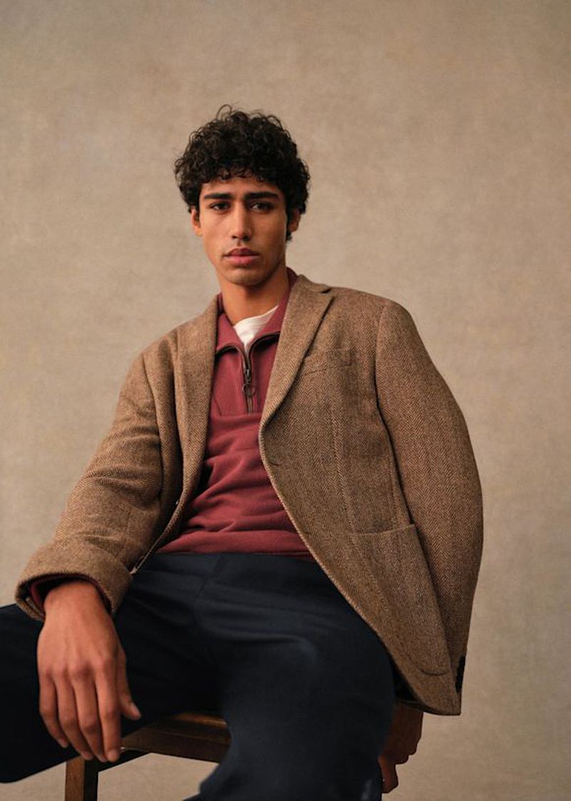 An adult male model with curly hair sits on a chair wearing a brown herringbone jacket and a burgundy polo shirt against a neutral backdrop.