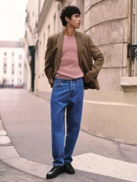 Man in a brown tweed blazer, pink knit shirt, and relaxed blue denim jeans standing on a city street sidewalk.
