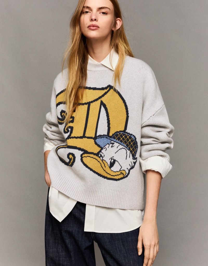 Woman wearing a grey sweater with a cartoon duck, paired with a white collared shirt and jeans, against a neutral grey backdrop.