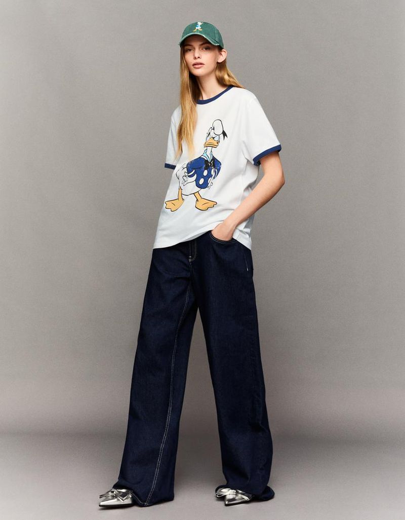 Adult woman poses in a white t-shirt with a Donald Duck cartoon, wide-legged jeans, green baseball cap, and silver shoes in a studio shot.