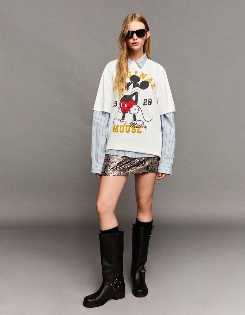 Fashion shot of a blonde woman wearing a Mickey Mouse t-shirt over a collared shirt with a sequin skirt and black boots in a studio.