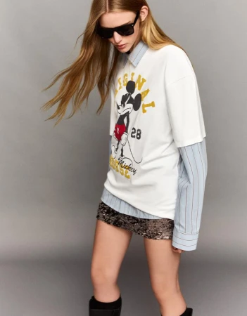 Young woman in a studio setting wearing a t-shirt with Mickey Mouse graphic and sequin skirt, styled with sunglasses and knee-high boots.