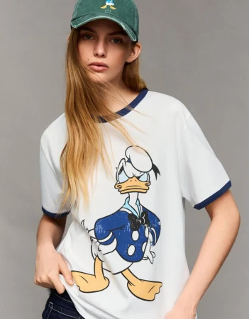 Female fashion model in studio wearing a white t-shirt with a Donald Duck cartoon graphic, paired with a green baseball cap.