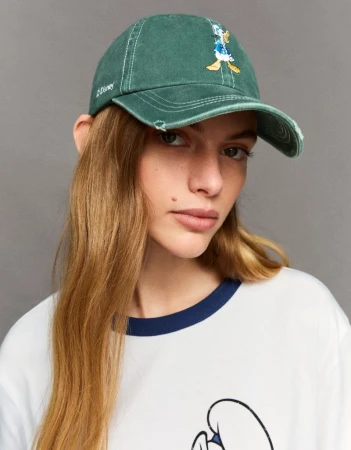 Woman in a green cap with Donald Duck graphic, wearing a white t-shirt with a blue-trimmed collar, against a gray background.