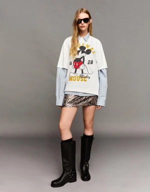 Model wearing a Mickey Mouse graphic tee over a striped collared shirt, sequin mini skirt, and black boots against a grey studio backdrop.