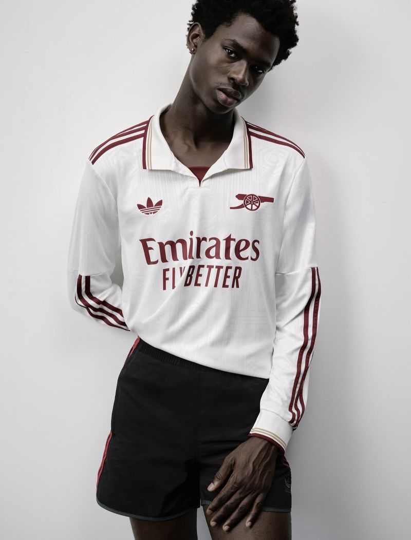 Athletic Black male model poses in a studio wearing a vintage white Arsenal soccer jersey and black shorts. Light, neutral studio background.