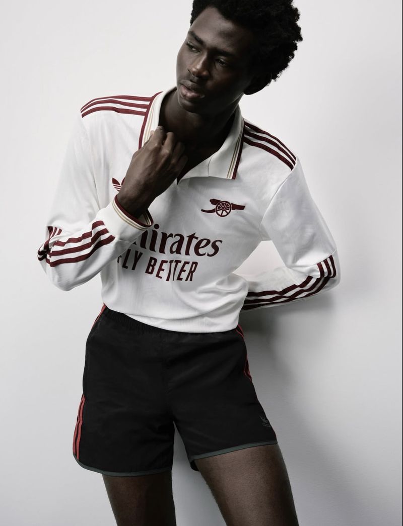 Athletic man poses in a white Arsenal soccer jersey with burgundy stripes and black shorts against a light grey background.