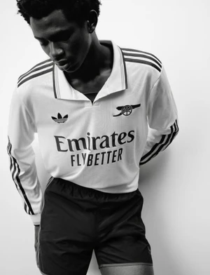 Model Willie Fidelis wearing a white long-sleeve Adidas Arsenal soccer jersey and dark shorts in a black and white studio portrait.