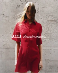 Model wearing a red button-down dress standing against a weathered concrete wall for Ami