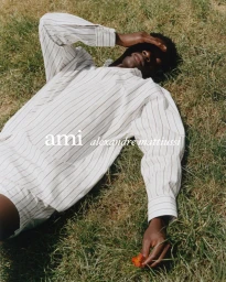 Model lying on grass wearing a white striped shirt holding a small red flower for the Ami Spring-Summer 25 campaign.