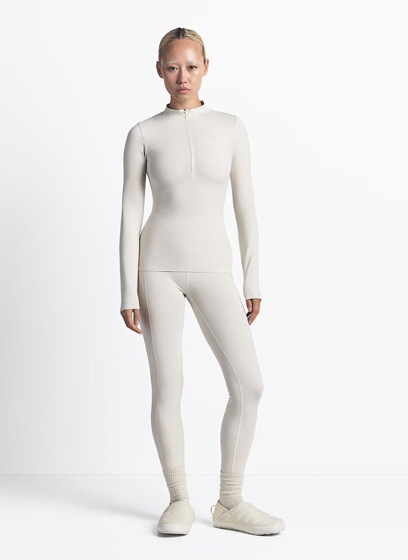 Full-body shot of a young adult female posing in a studio, wearing a light-colored long-sleeve top, leggings, and matching shoes.