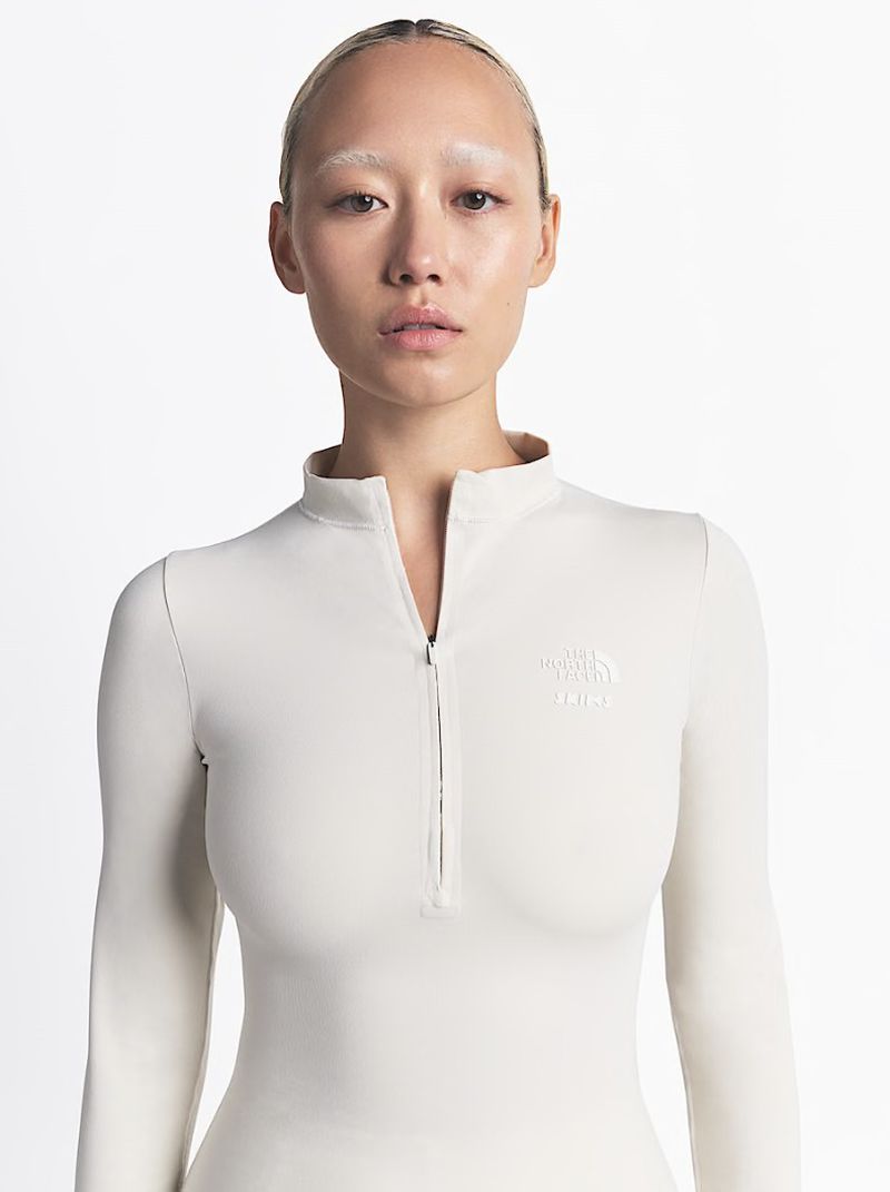 Close-up studio shot of an adult woman wearing a form-fitting, long-sleeve top with a partial zipper and featuring white eyebrows.