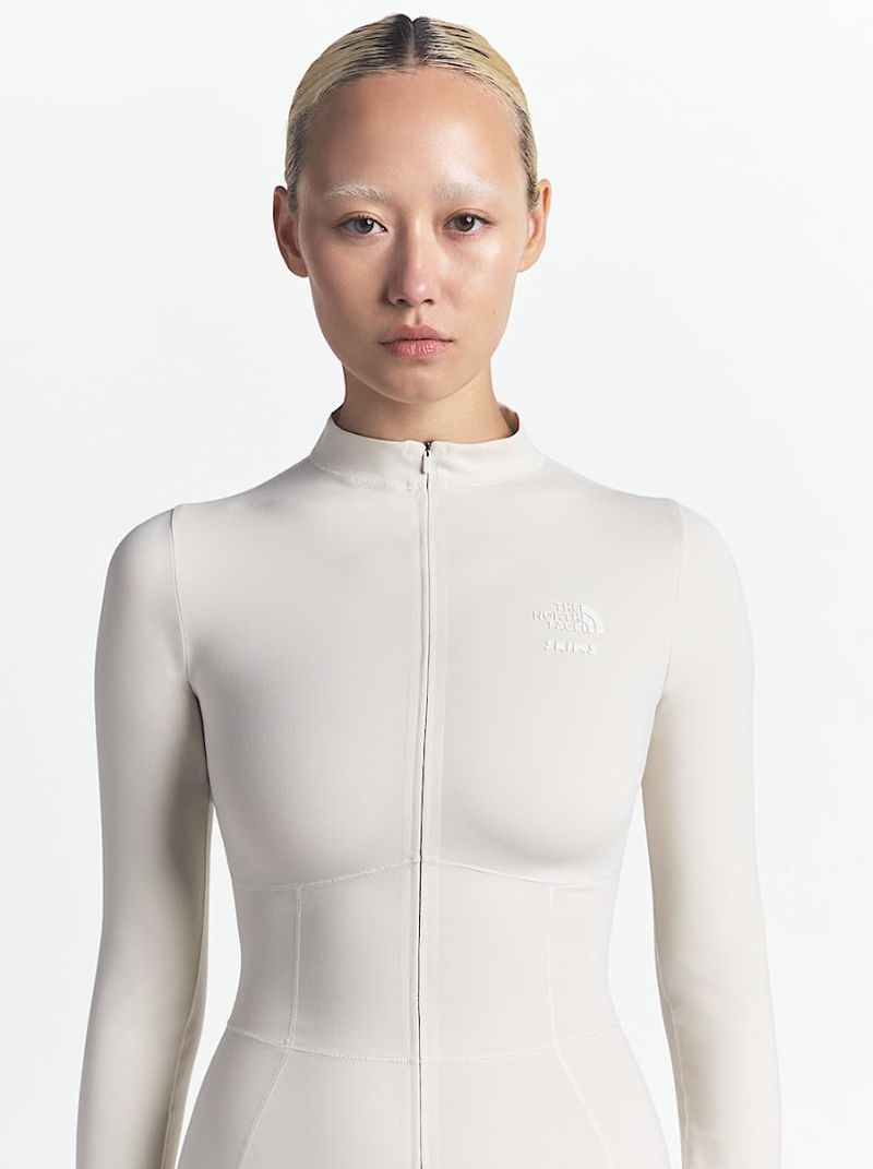 Close-up studio shot of an adult woman with pale skin and light hair, wearing a light beige long-sleeve bodysuit against a white backdrop.