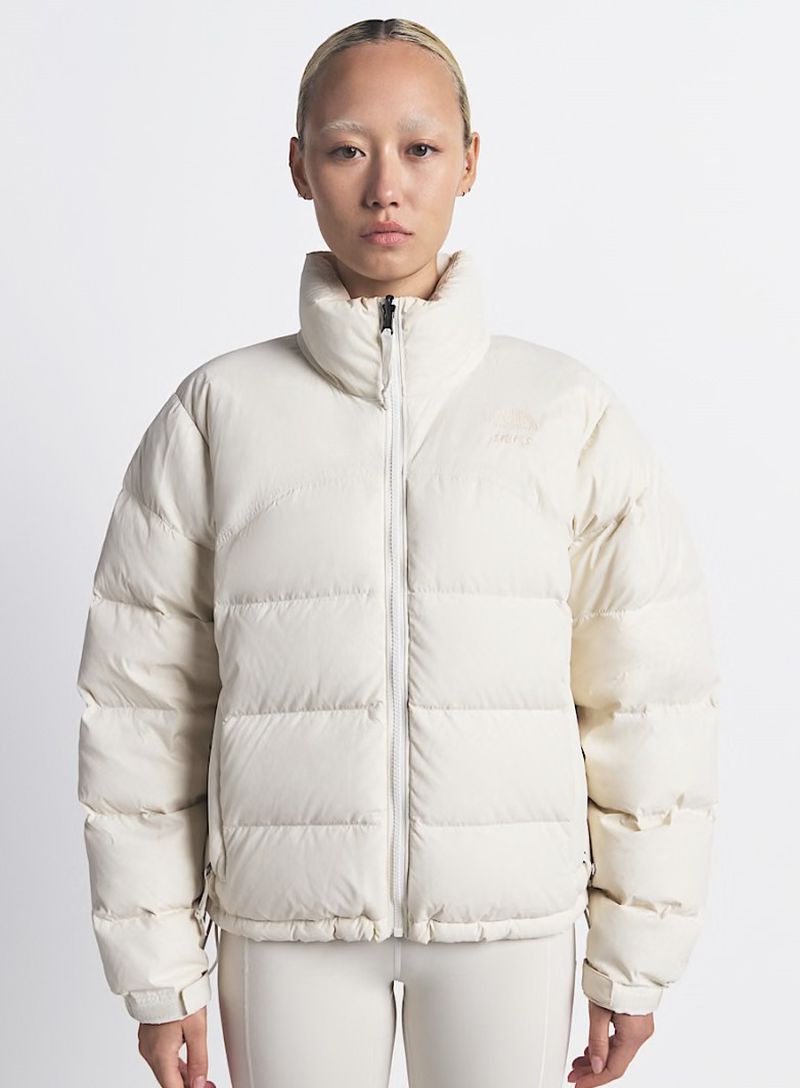 A person wears a cream-colored puffer jacket and matching pants in a studio shot, showcasing the garment's design and fit on a neutral backdrop.