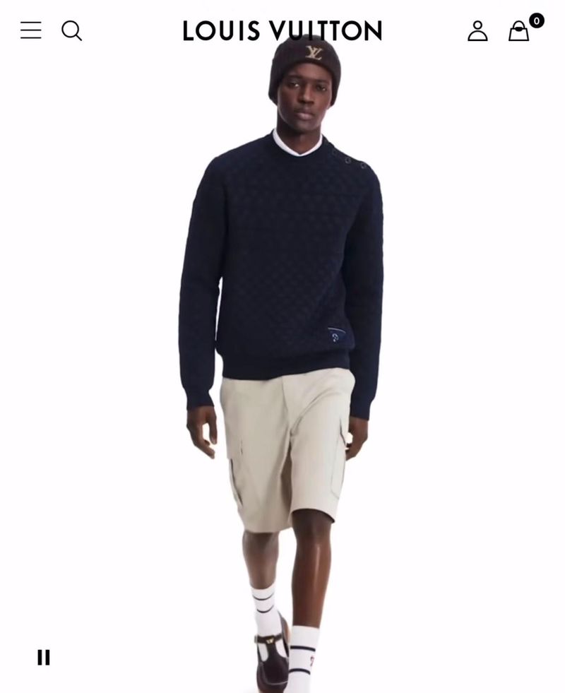 A dark-skinned male model wears a dark blue sweater, khaki shorts, beanie, and Mary Jane shoes by Louis Vuitton in a studio shot.