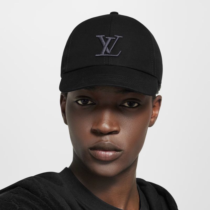 Close-up of an African-American adult female wearing a black baseball cap with the Louis Vuitton logo and a black shirt against a neutral background.
