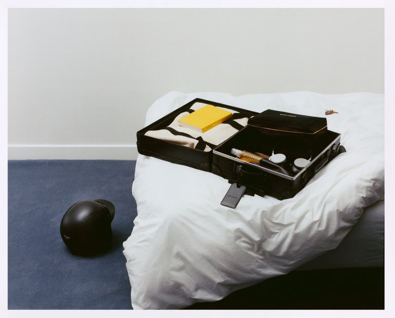 Open black suitcase with toiletries and yellow book on a white bed, with a black cap on a blue carpet in a white-walled room.