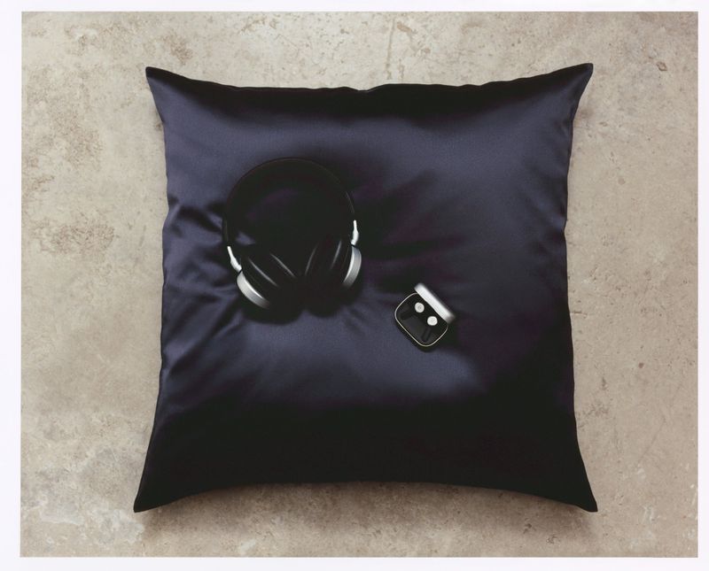 Overhead shot of a pillow with headphones and a smart watch on it, with a concrete-like surface visible in the background.