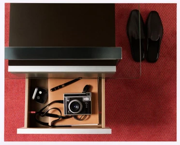 Top-down view of an open desk drawer containing a camera, pen, and notebook beside black loafers on a red carpet.