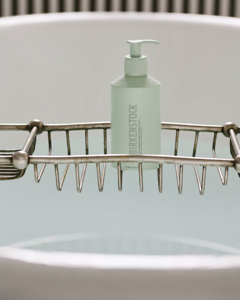 Birkenstock cleansing milk and body wash bottle with dispenser on metal rack over filled bathtub, white bathroom interior.
