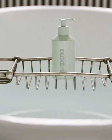 Birkenstock cleansing milk and body wash bottle with dispenser on metal rack over filled bathtub, white bathroom interior.
