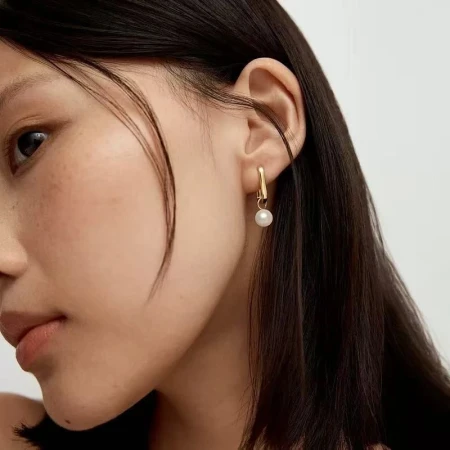 Close-up profile of an East Asian woman wearing a gold hoop earring with a dangling white pearl against a light background.