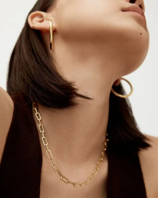 Close-up of a woman wearing gold jewelry including a chain necklace and large hoop earrings, against a neutral background.