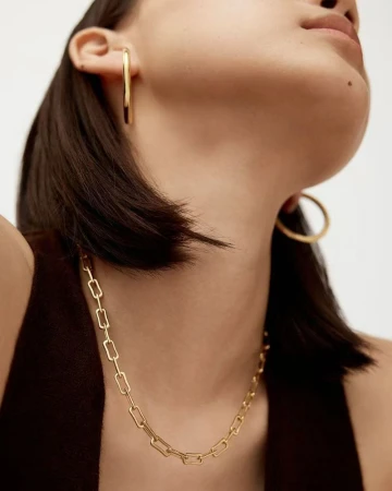 Close-up of a woman wearing gold jewelry including a chain necklace and large hoop earrings, against a neutral background.